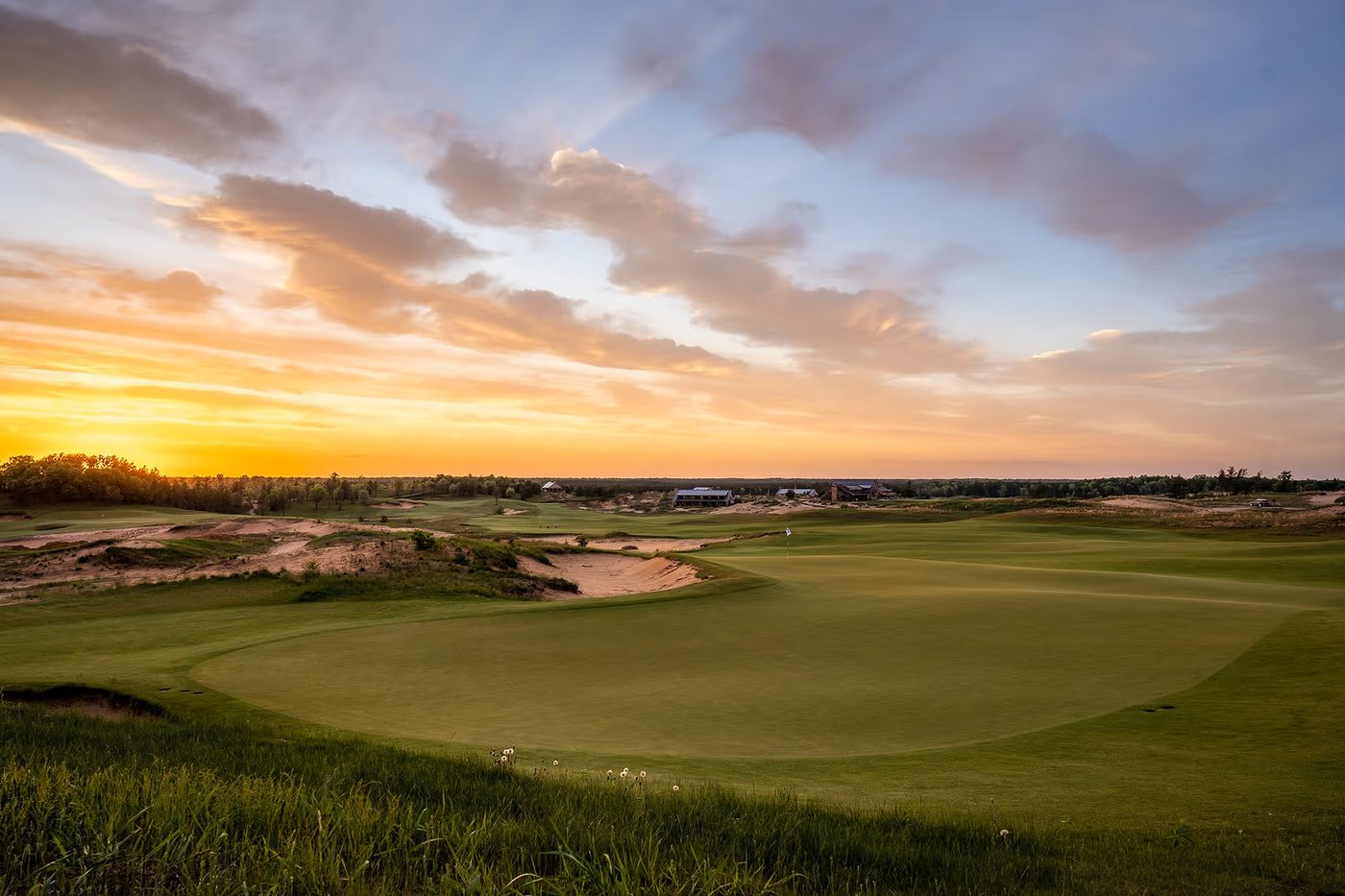 A wide green with sand faces under a streaked orange sunset at Sand Valley