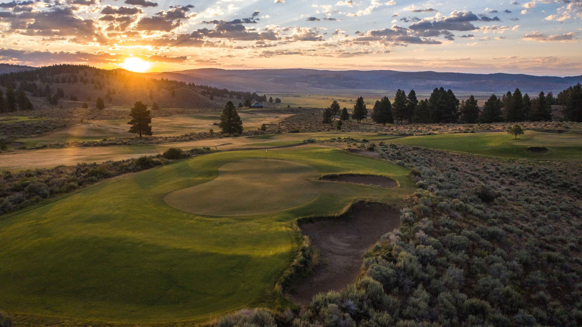 Sun setting over the valley behind a bunkered green framed by sagebrush and pines at Silvies Valley Ranch