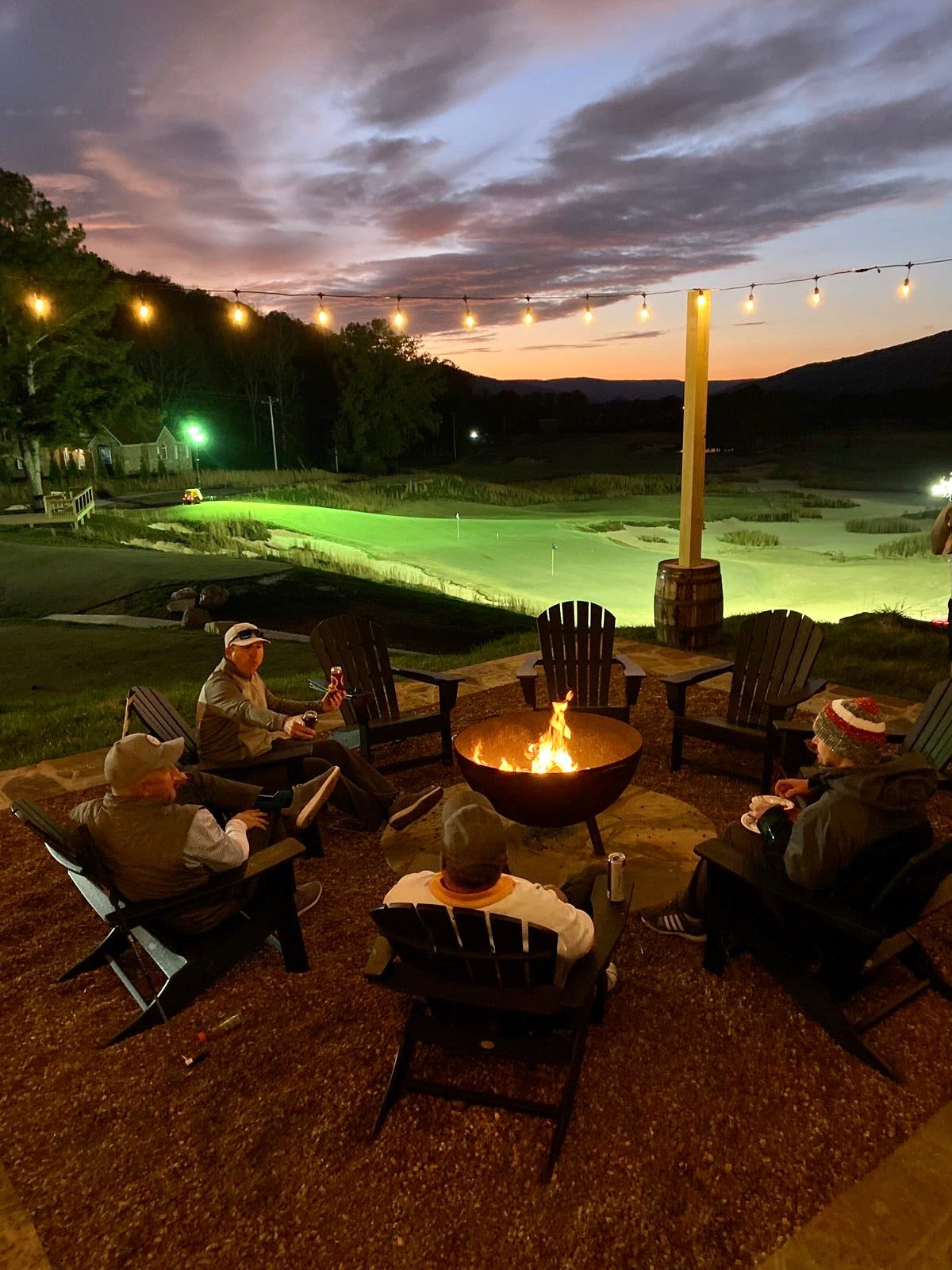 Golfers in Adirondack chairs around a fire pit under string lights at Sweetens Cove