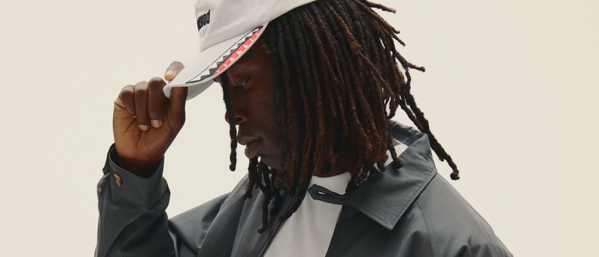 From the Metalwood FW26 Pt 1 lookbook: a man in the steel Raglan Sleeve Blouson tipping the brim of a white Bomber Dad Hat with a shark mouth on it