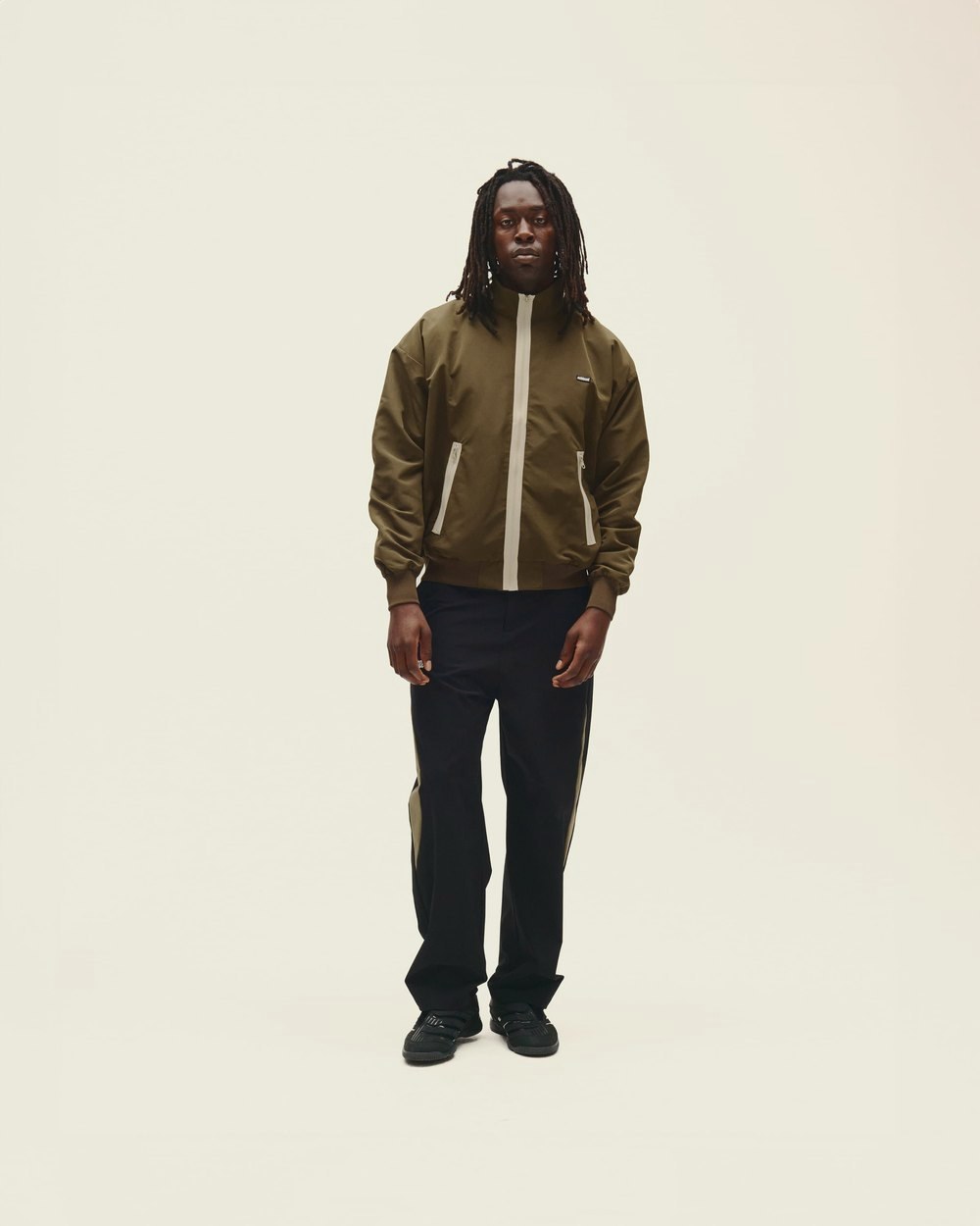 A man in the brown Contrast Zip Windbreaker and black pants standing against a white wall
