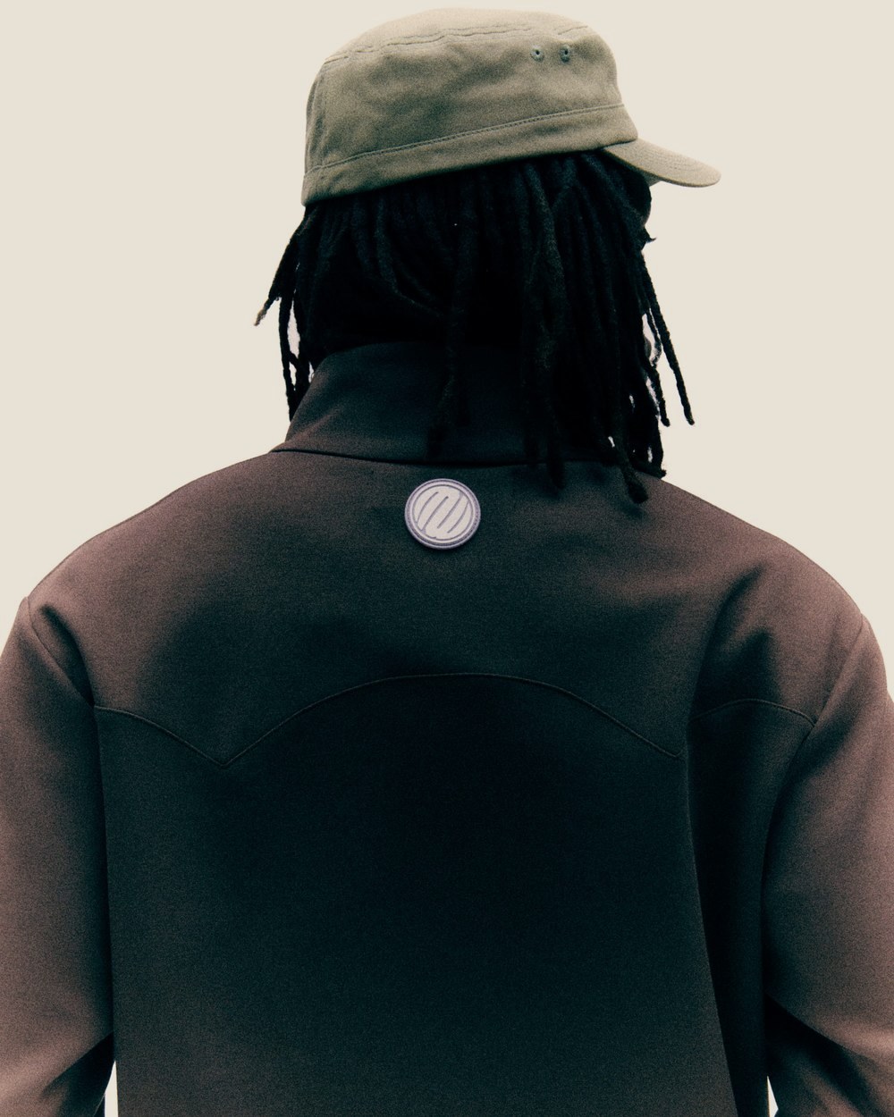 A man seen from behind in a tan cadet cap and the brown Western Yolk Track Jacket with the circular M badge
