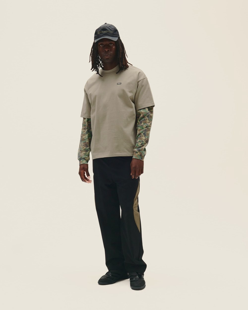 A man in a tan tee over a camo long sleeve, with black pants and a dark cap