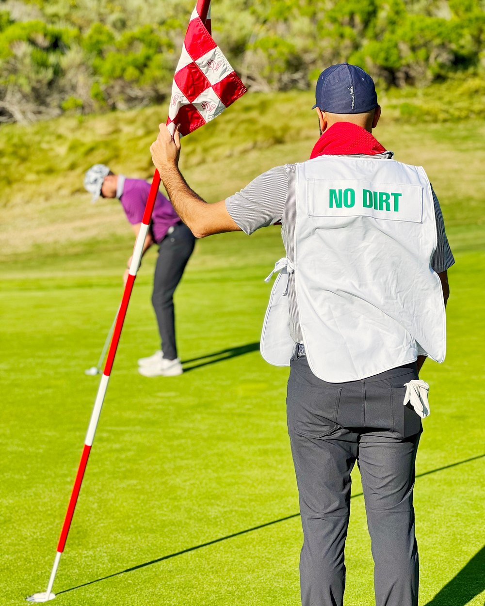 A caddie in a white No Dirt bib holding the flagstick on a green while a golfer putts