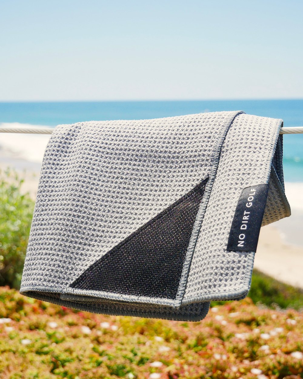 A gray No Dirt Classic towel hanging on a rail above a beach