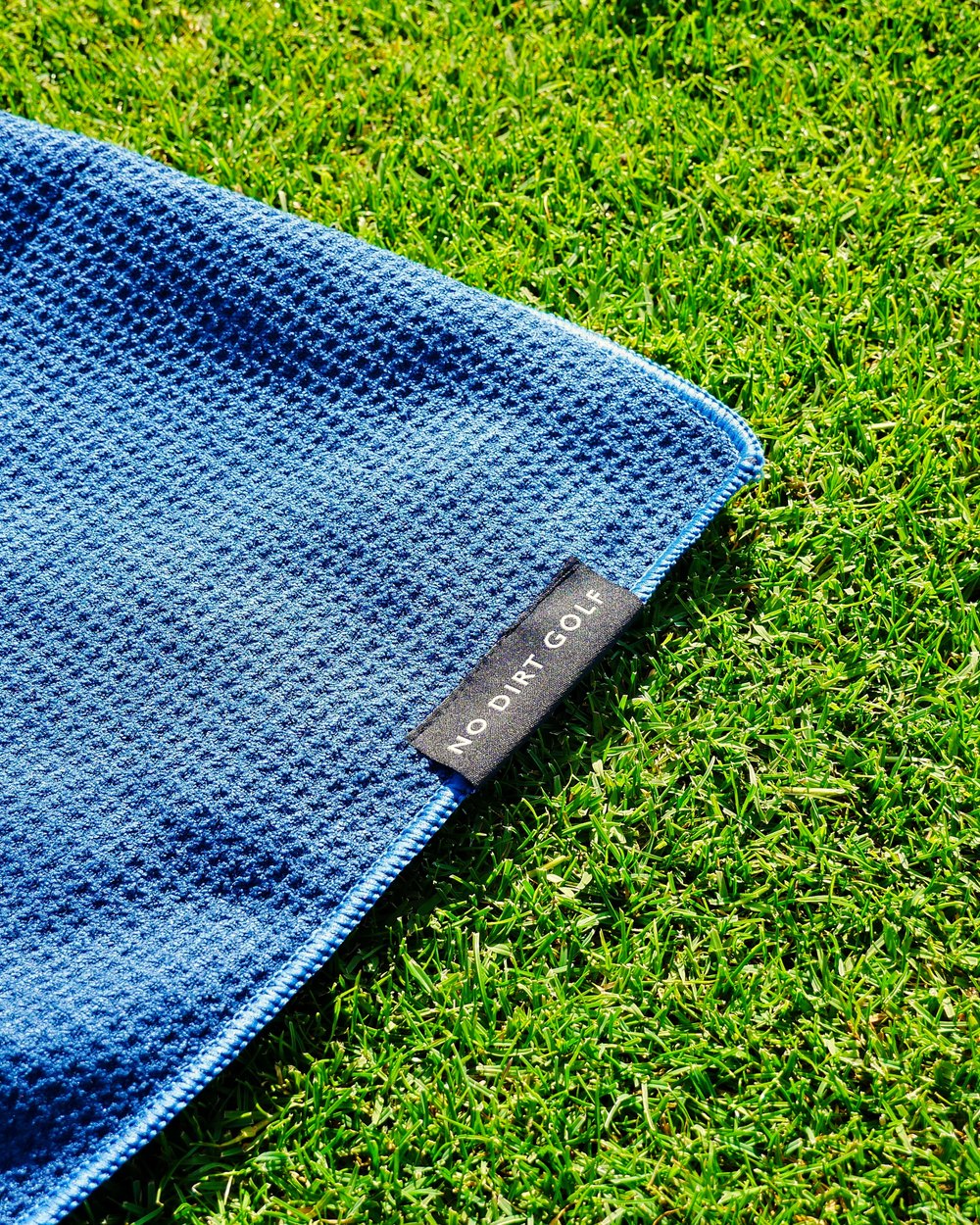 A blue No Dirt waffle towel with its woven label lying on grass