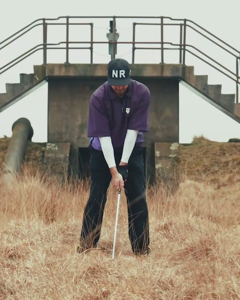 A golfer in an NR cap and purple Wind Shirt addressing a ball in long grass