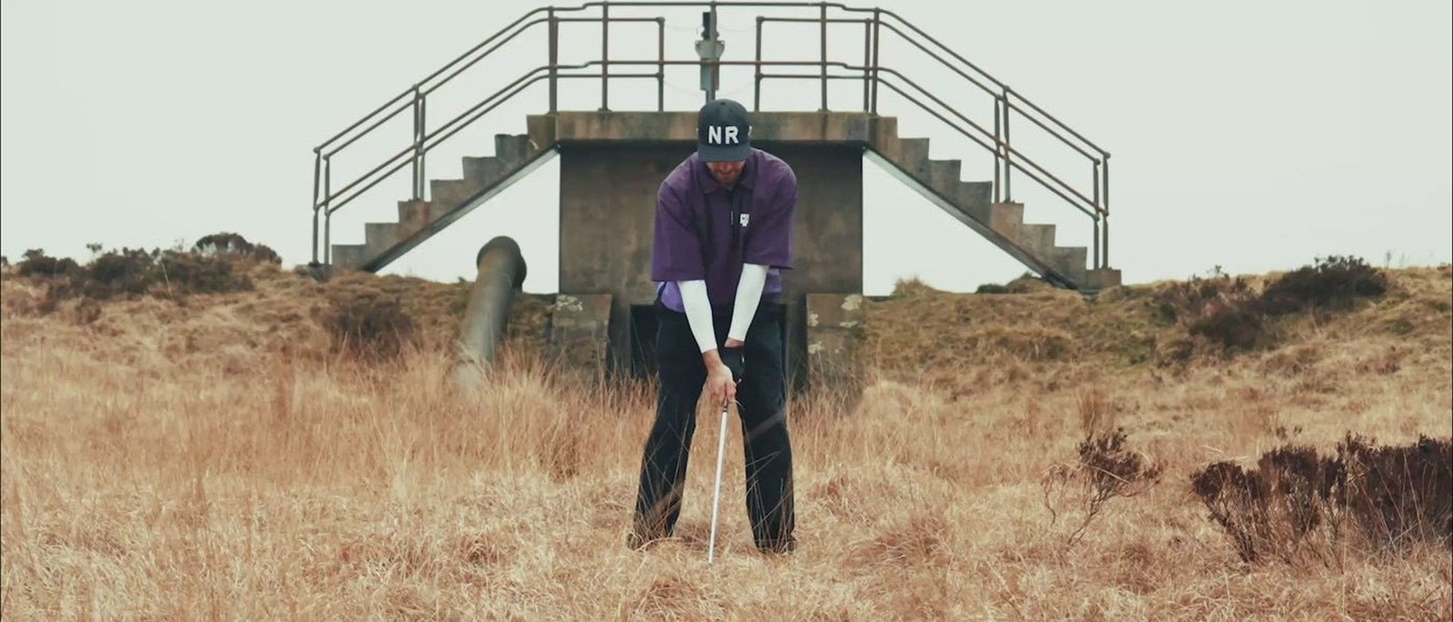 A golfer in an NR cap and purple No Return Club Wind Shirt addressing a ball in long moorland grass in front of a concrete structure with steel stairs