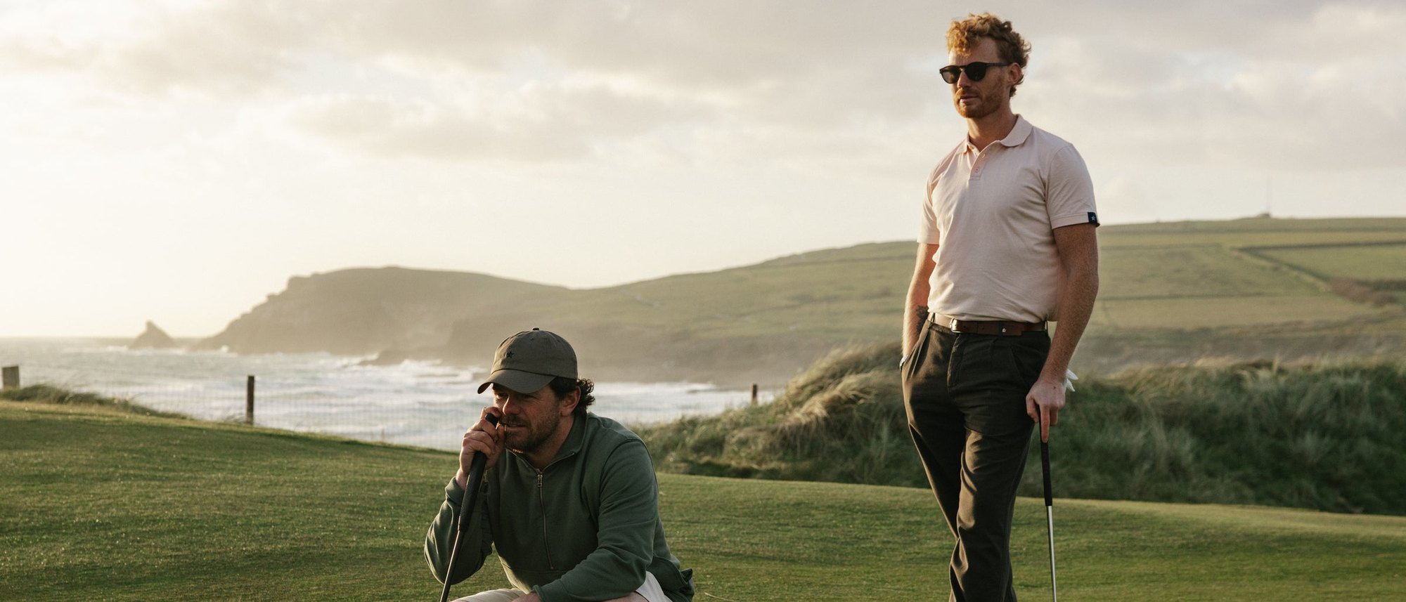 Two golfers on a clifftop links course above the sea, one crouching to read a putt, from Sounder’s own shoot