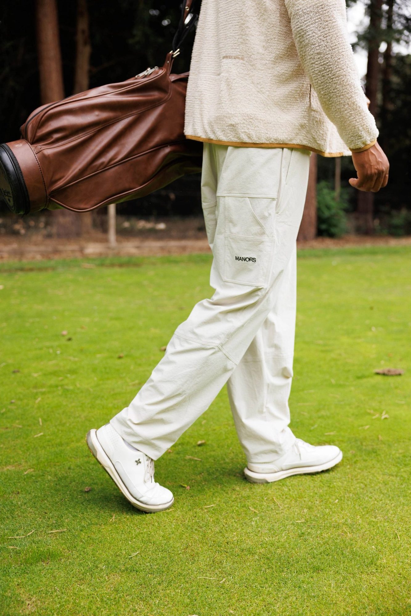 A golfer walking across a green in ivory Manors Greenskeeper trousers and white trainers