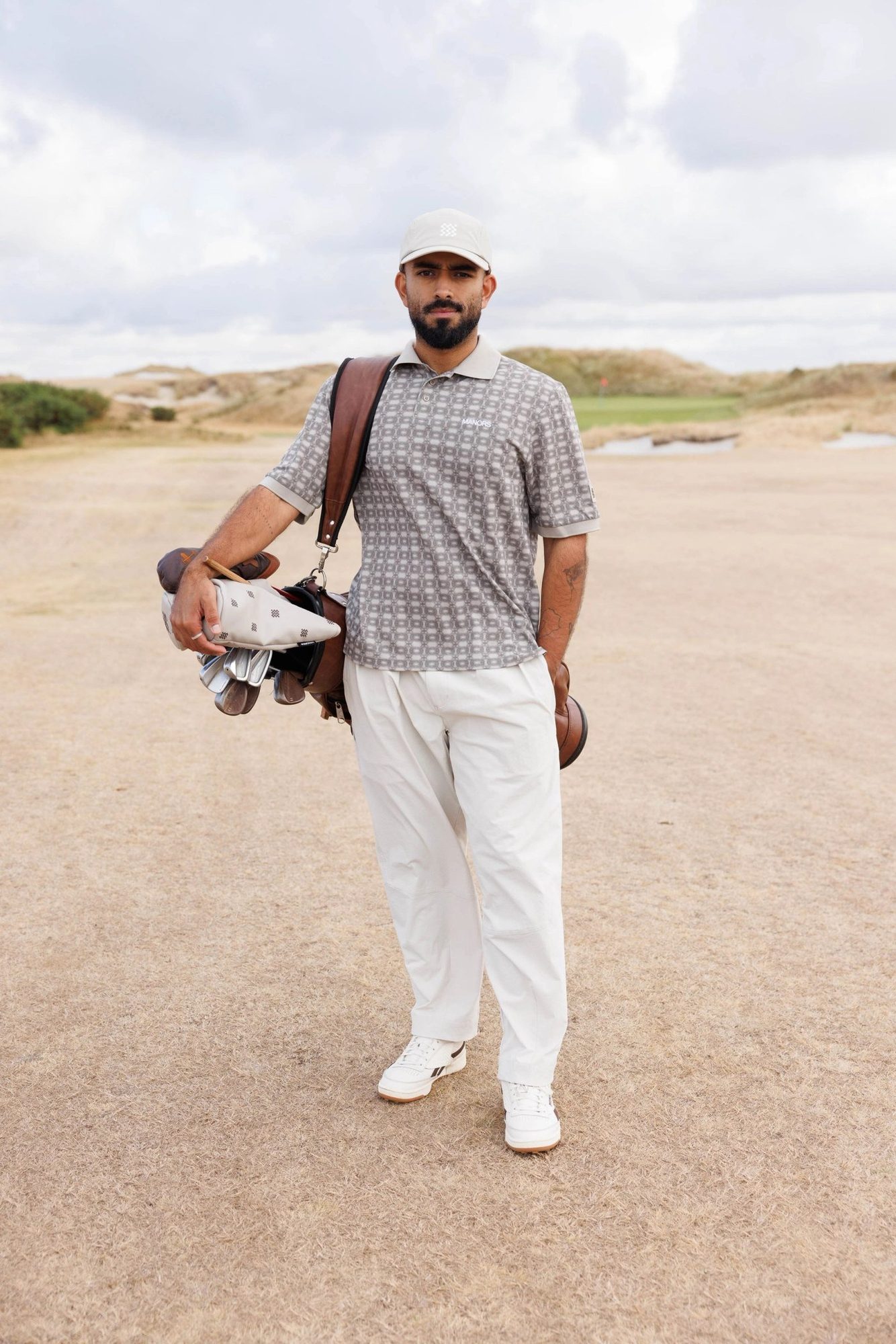 A golfer standing on a sandy path with his bag in ivory Greenskeeper trousers