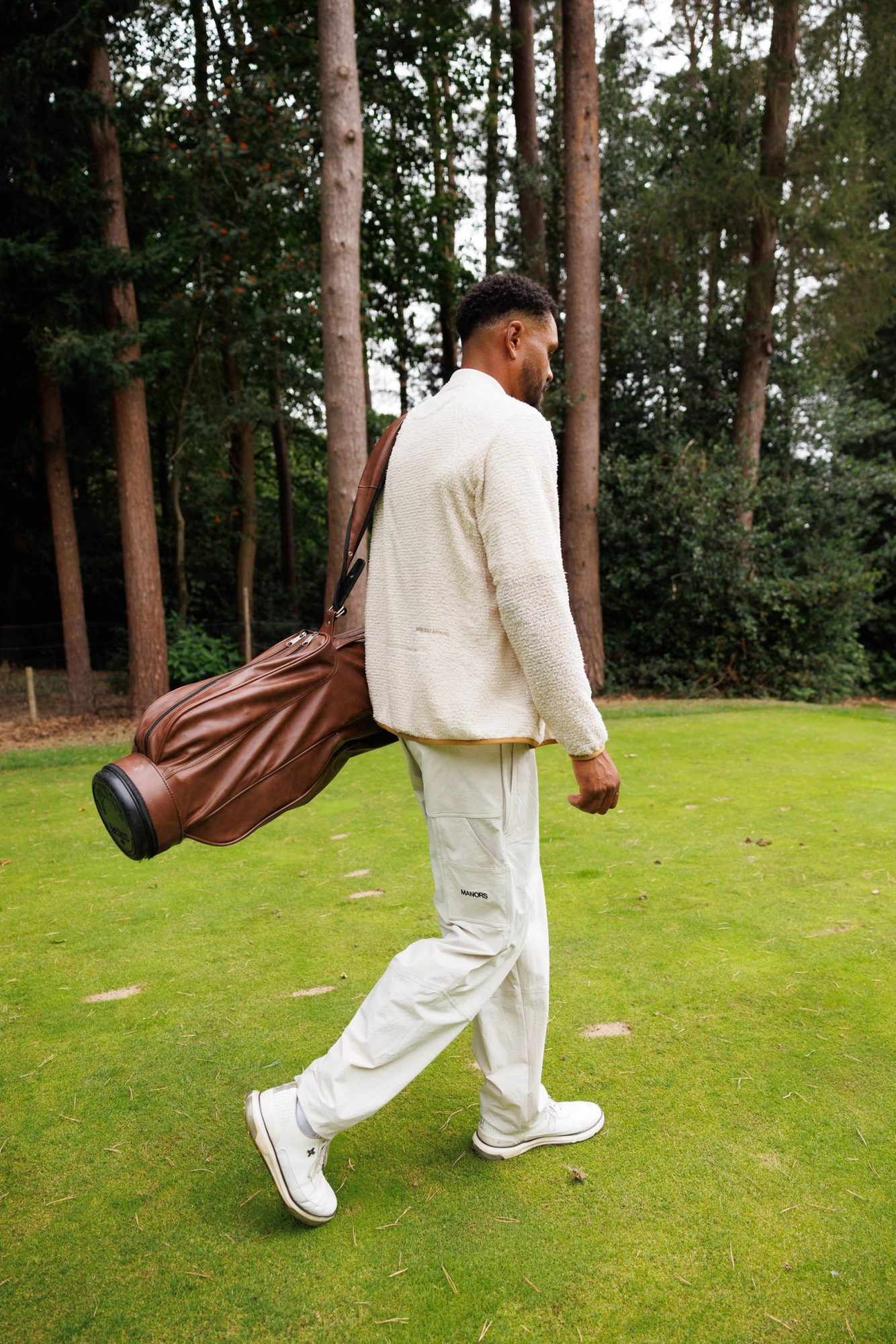 A golfer carrying a leather bag through a pine-lined fairway in ivory Greenskeeper trousers