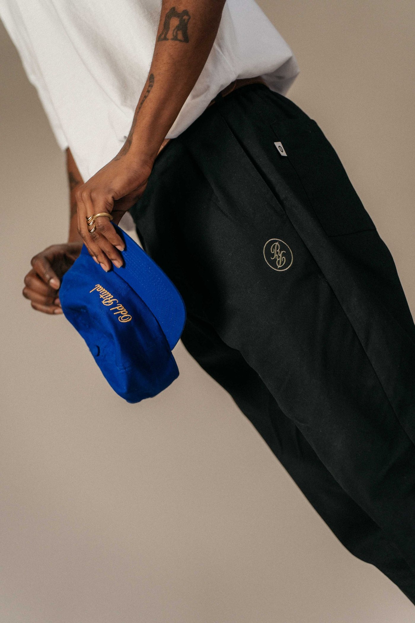 Close detail of the black Pleated Daily Trouser showing the embroidered RR monogram on the thigh, with a blue Odd Ritual cap in hand