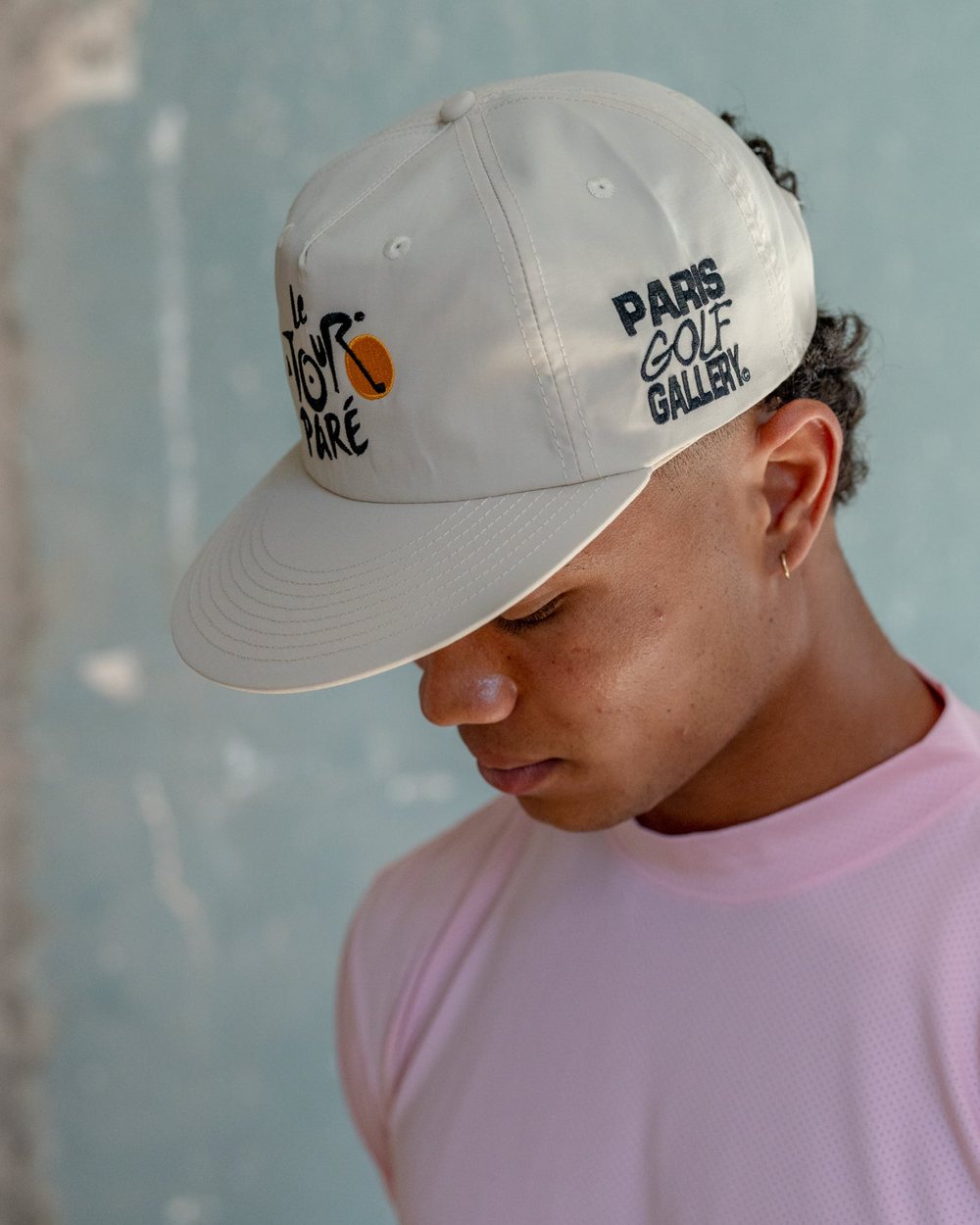 A man in a pink long sleeve and the cream Paris Golf Gallery x Paré cap looking down