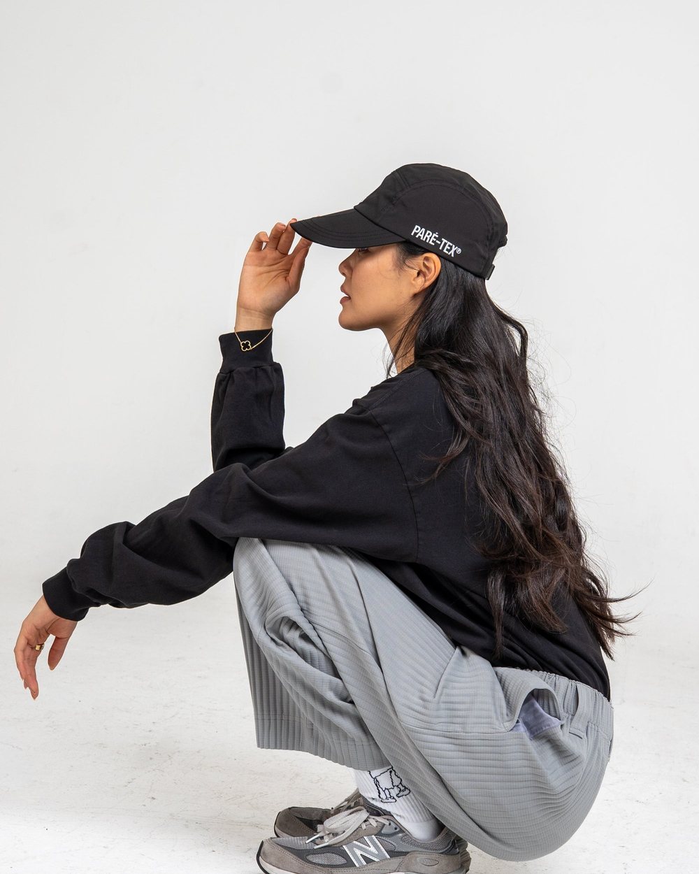 A woman in a black Manila long sleeve, grey Plissé pants and a black cap crouching