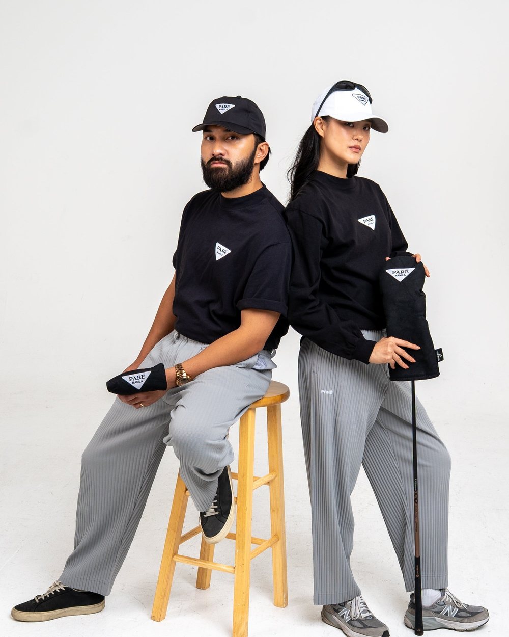 Two people in black Manila tees and grey Plissé pants on stools holding the Bluetross driver and putter covers
