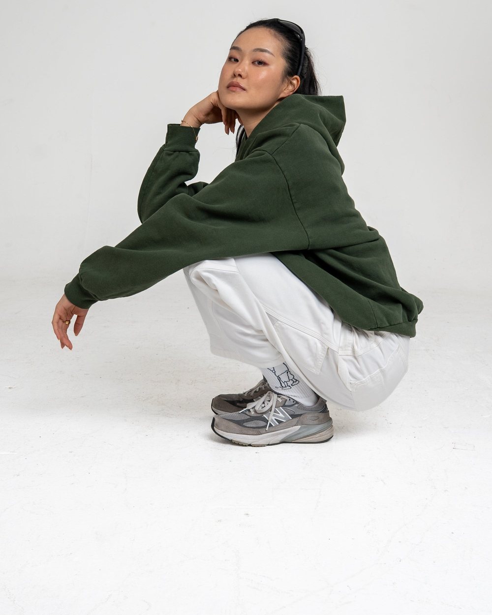 A woman in the green Gulay hoodie and grey Plissé pants crouching with a hand on her chin