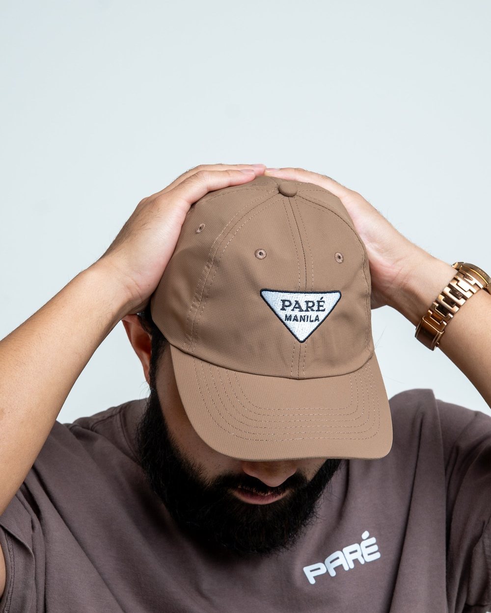 A man in a brown tee pulling down the walnut Imperial Manila cap