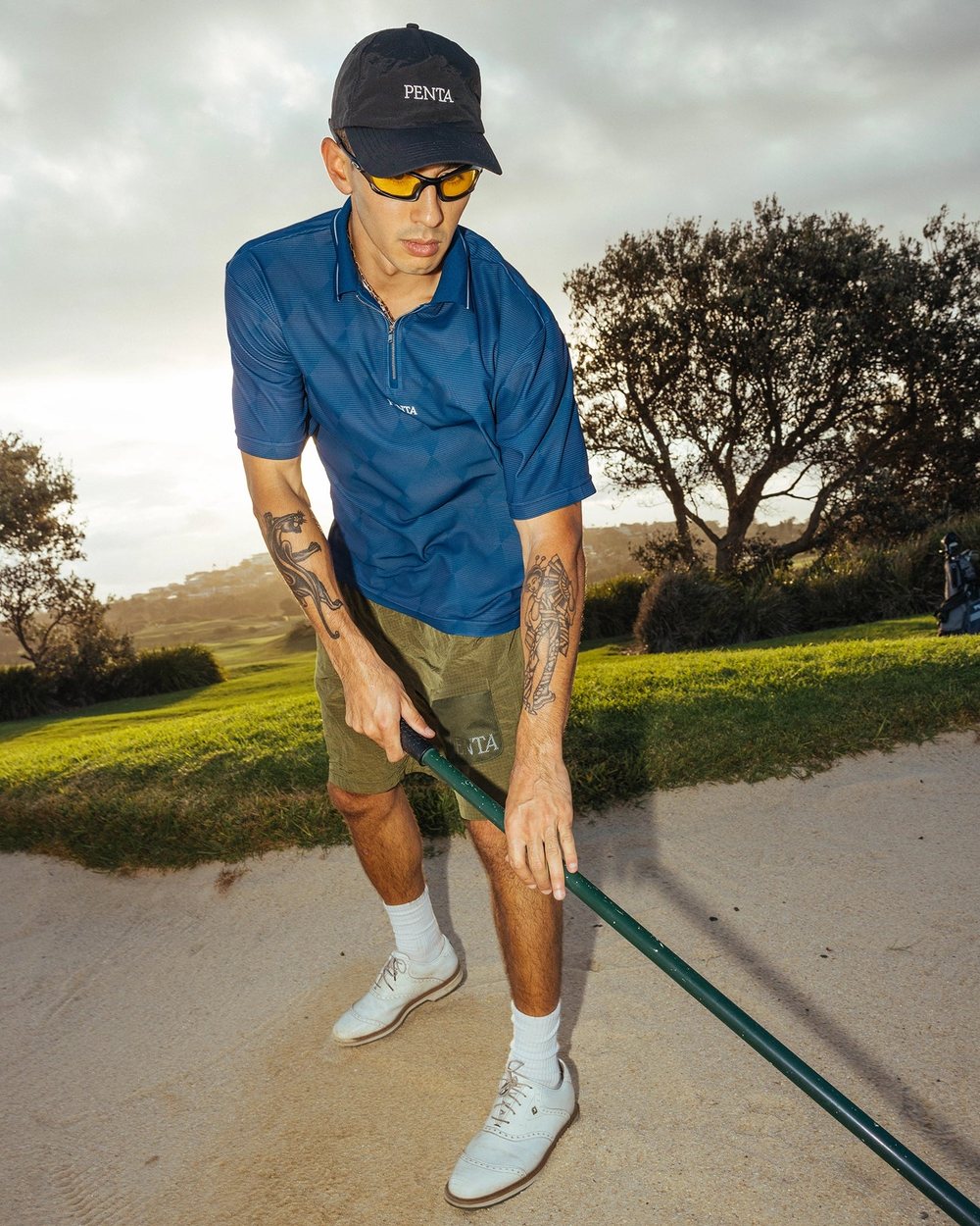 A golfer in moss Hazard Shorts and a navy cap playing out of a bunker