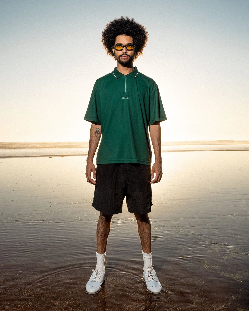 A man in the green Penta zip polo and black shorts standing on a beach at sunset