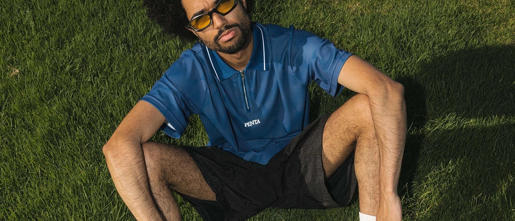 A man in orange-tinted sunglasses and the navy Penta Optical zip polo sitting on the grass on a sunny day