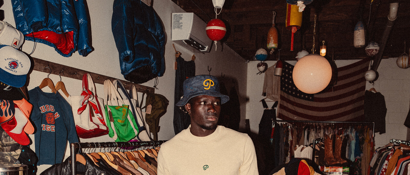 Public Drip FW26 campaign shot — a model in the indigo denim bucket hat and cream crewneck inside a dimly lit vintage clothing shop, racks and hanging lamps behind