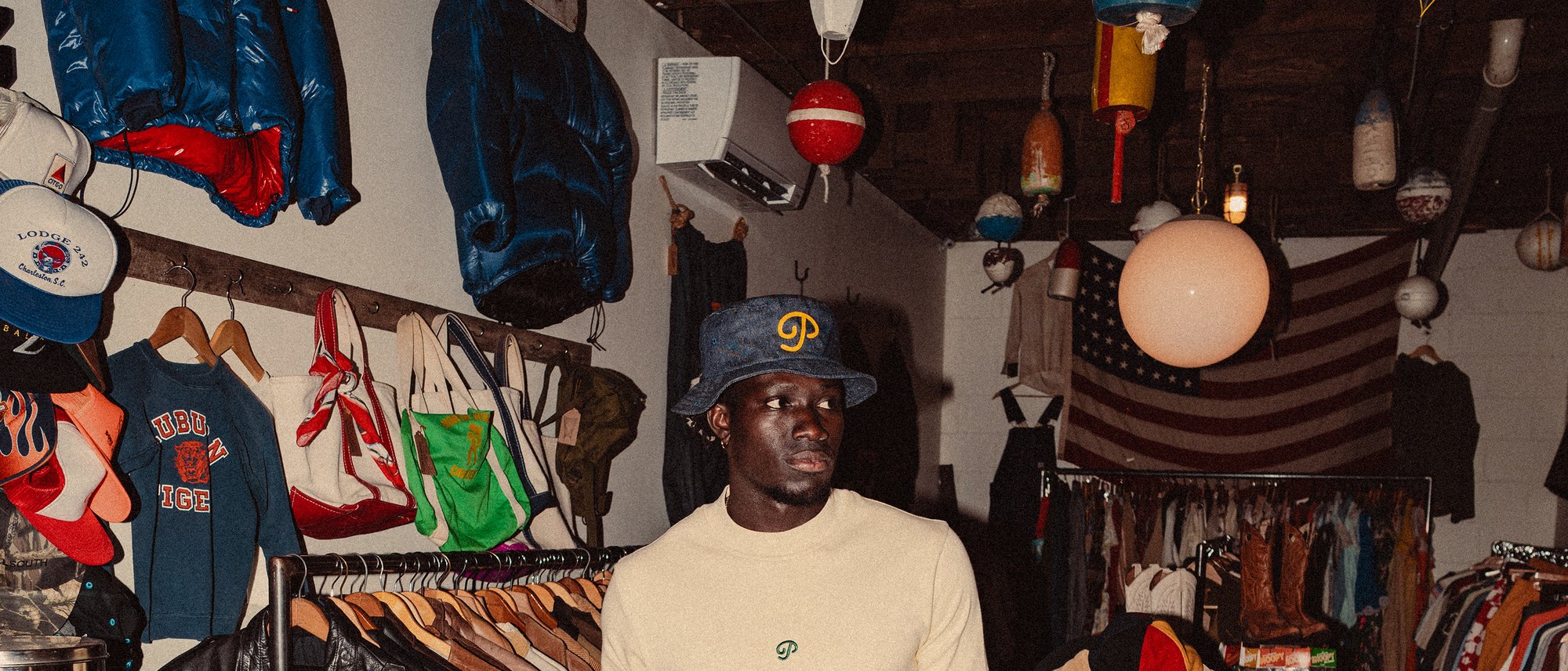 A man in the denim P script bucket and a cream long-sleeve in a vintage clothing shop, from Public Drip’s Nightshift campaign
