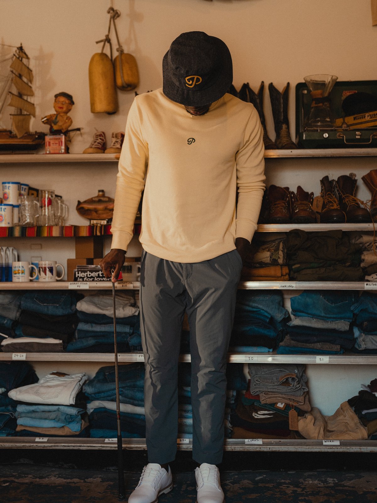 A man in a cream knit, grey trousers and a P script cap in a vintage shop