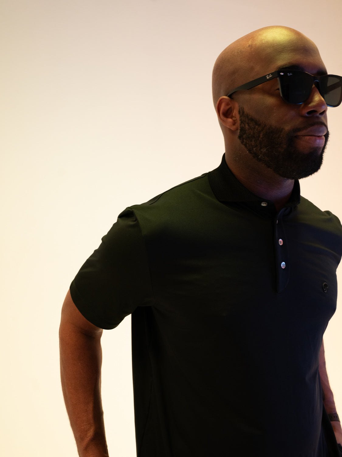 A man in sunglasses and the black tonal polo against a plain backdrop