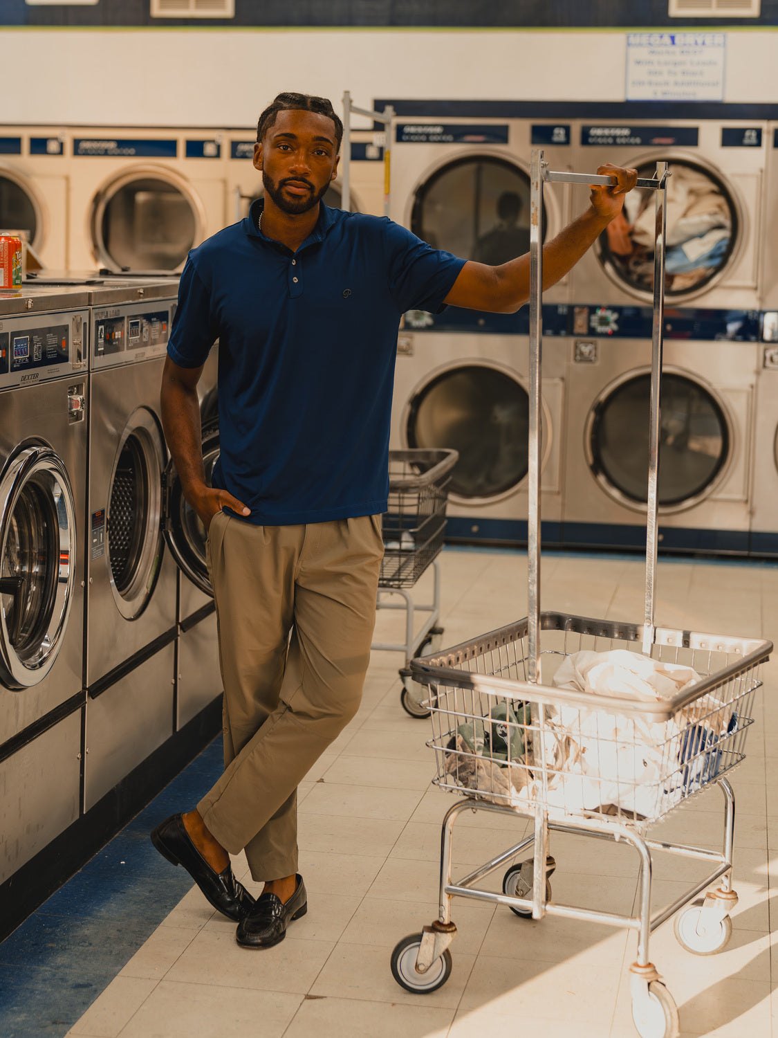 A man in the navy tonal polo and sand pants leaning on a laundromat cart