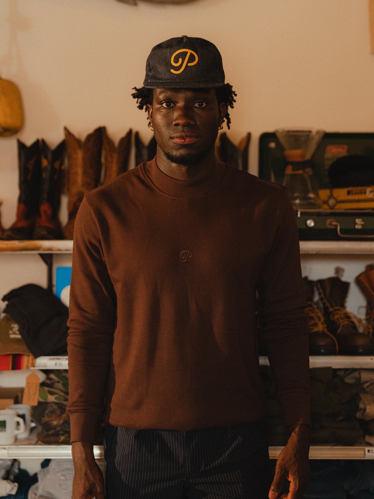 A man in a coffee knit and P script cap in front of shelves of vintage boots