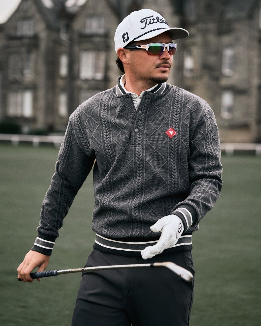 A golfer in a grey Macade cable-knit quarter-zip holding an iron in front of a stone clubhouse