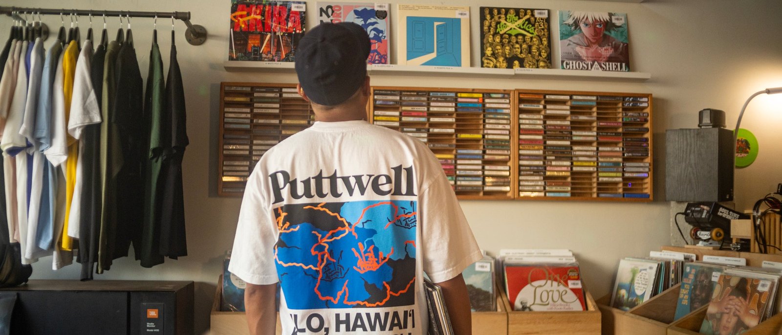 A man in a Puttwell tee, the Sig Zane back print facing out, browsing tapes and records in a Hawaii record store
