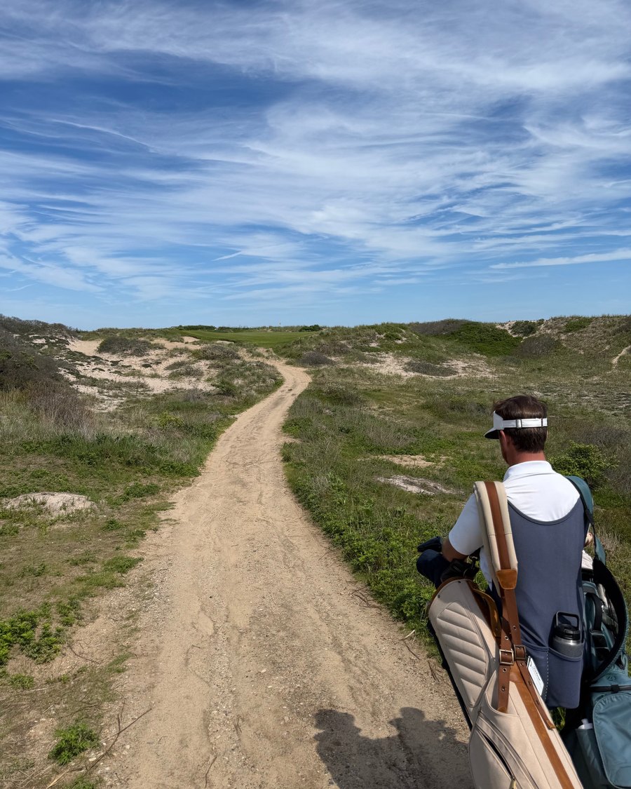 A golfer carrying a bag down a sandy path through the dunes