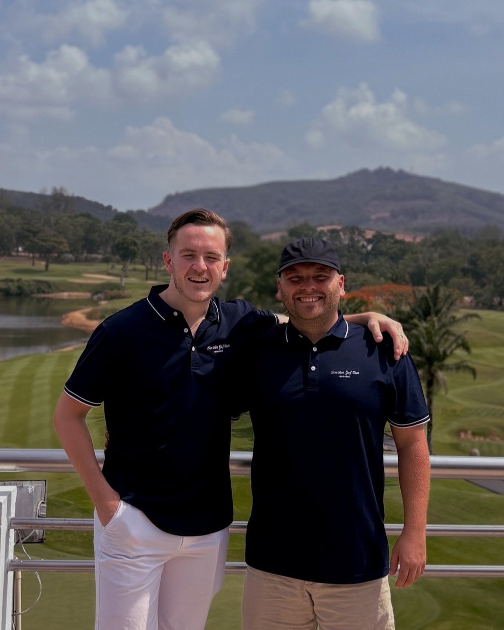 Two Dim Sum Golf Club members in navy polos on a terrace above a golf course and lake, with hills behind