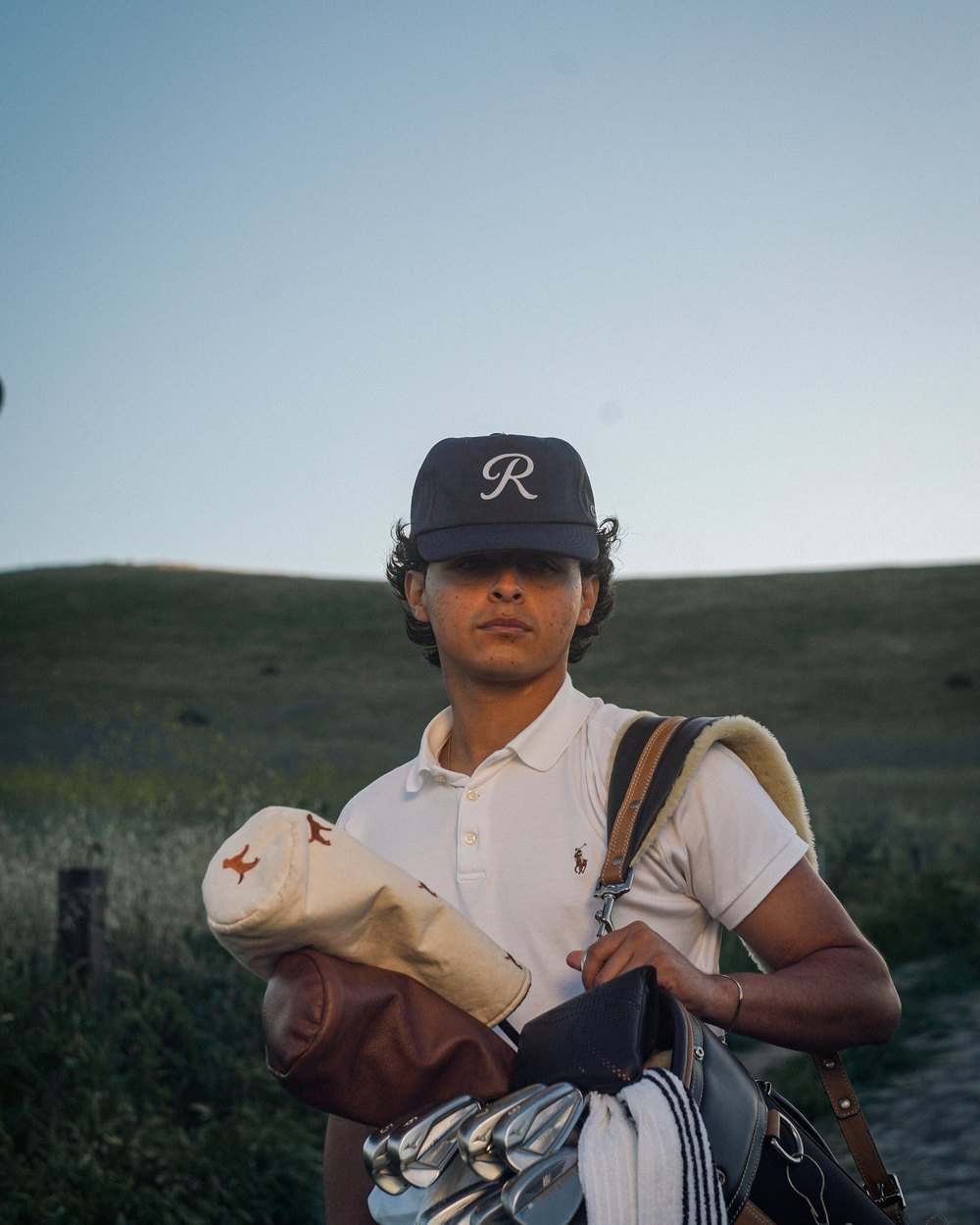 A young golfer in a navy R hat carrying a bag through long grass at dusk