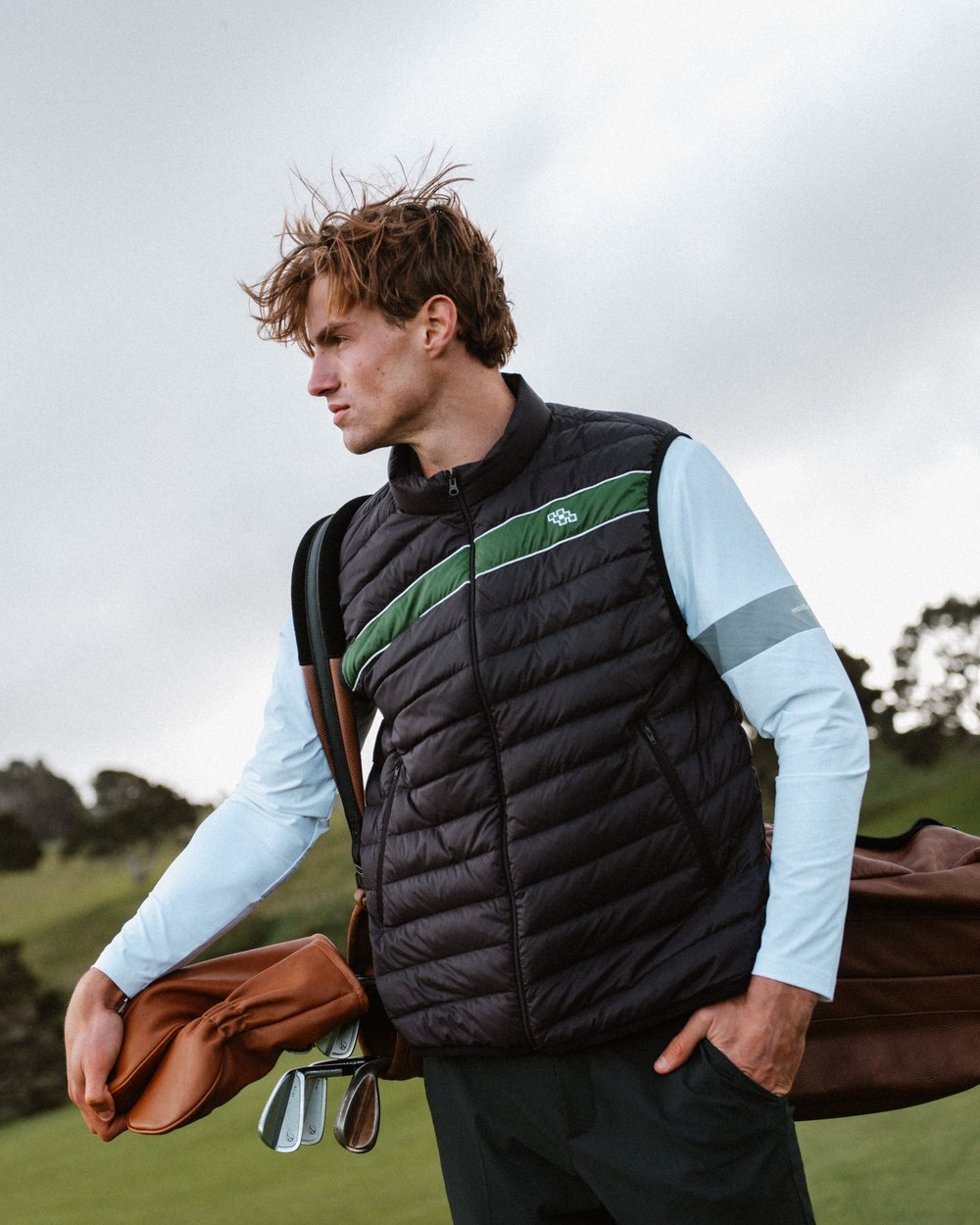 A golfer in a black puffer vest with a green stripe over a light blue shirt, carrying a bag on a course