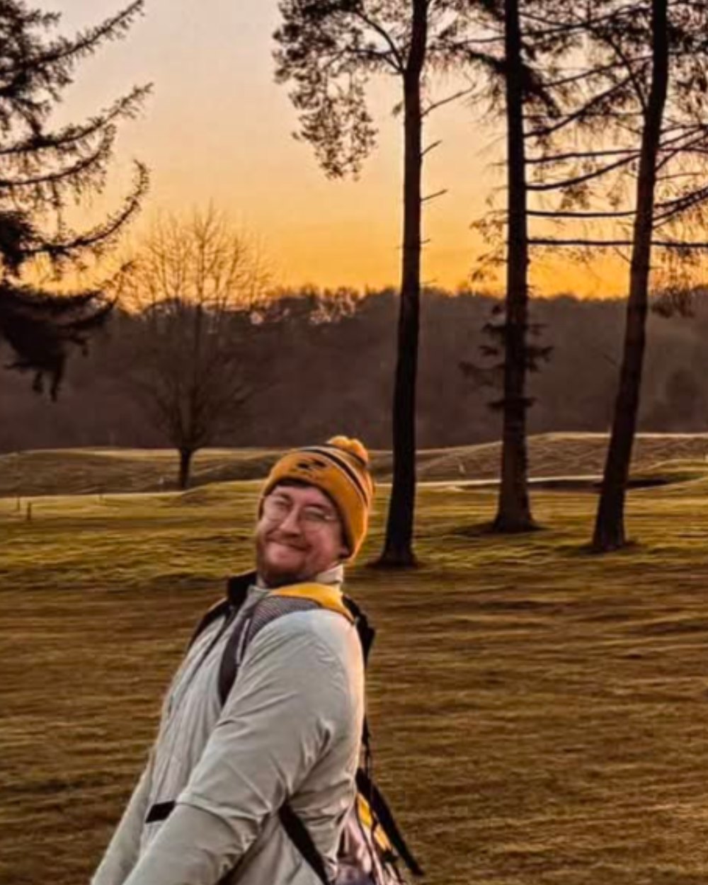 A smiling golfer in a beanie carrying his bag across a fairway at sunset