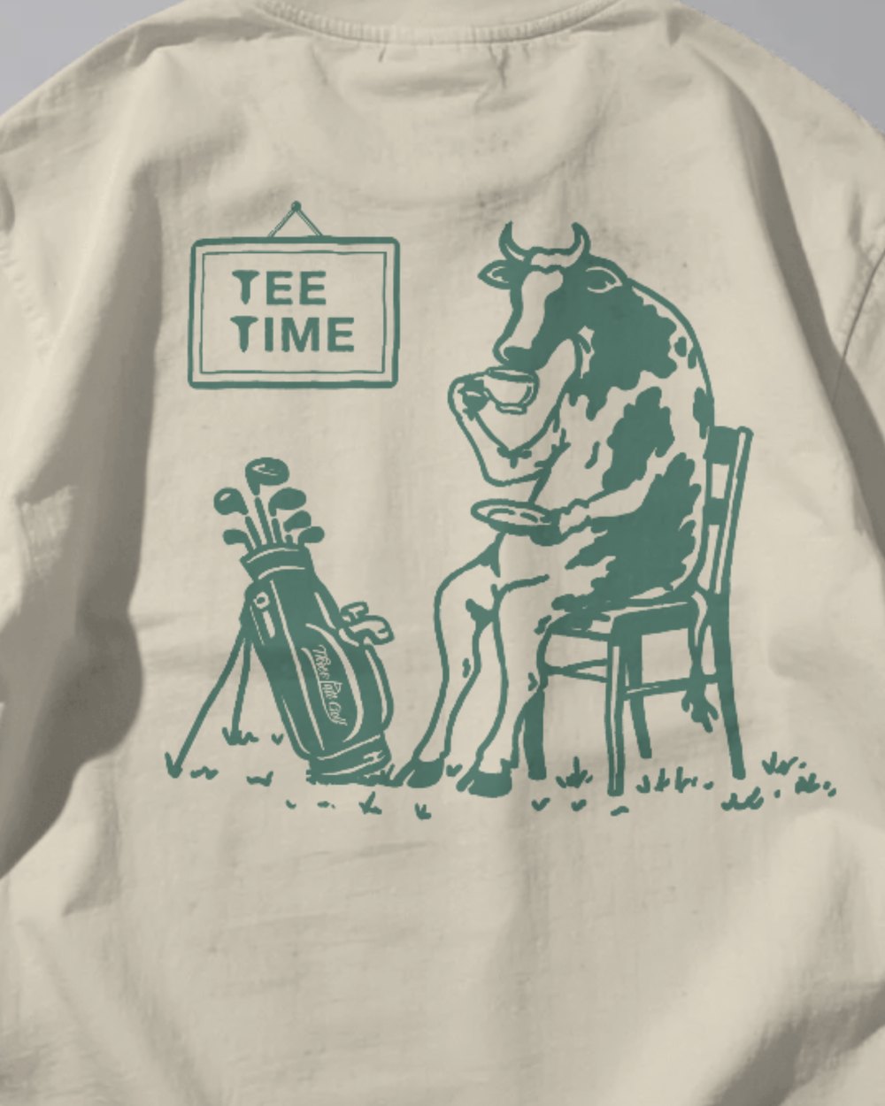 The Tee Time print on the back of a cream Three Putt tee: a cow on a chair with a cup of tea beside a golf bag