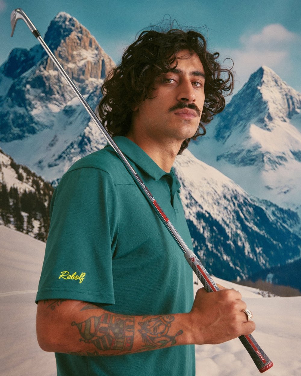A model in a green Rebolf polo holding a club over his shoulder in front of a snowy mountain backdrop