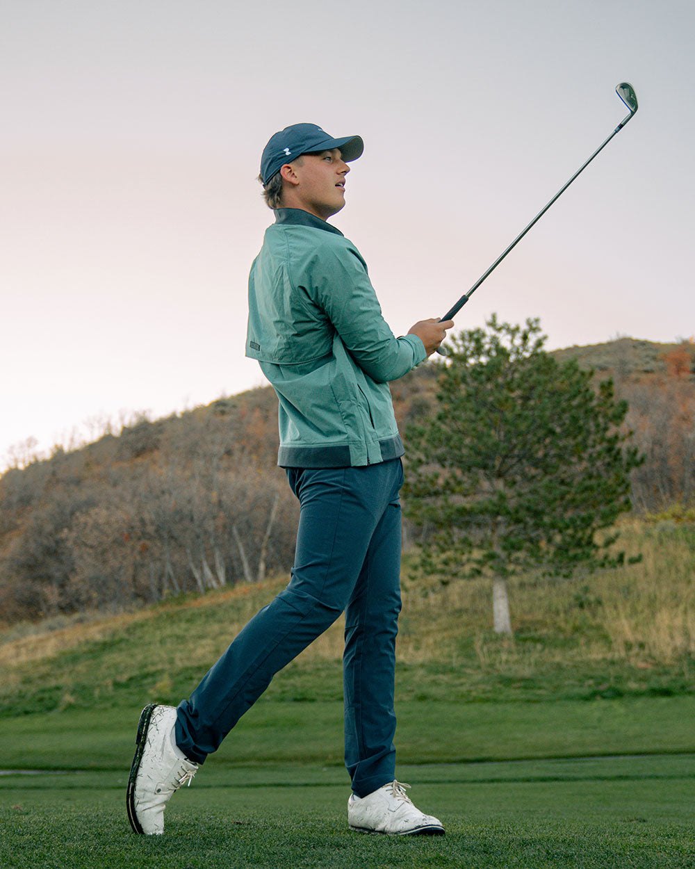 A golfer finishing a swing in navy pants and a green layer