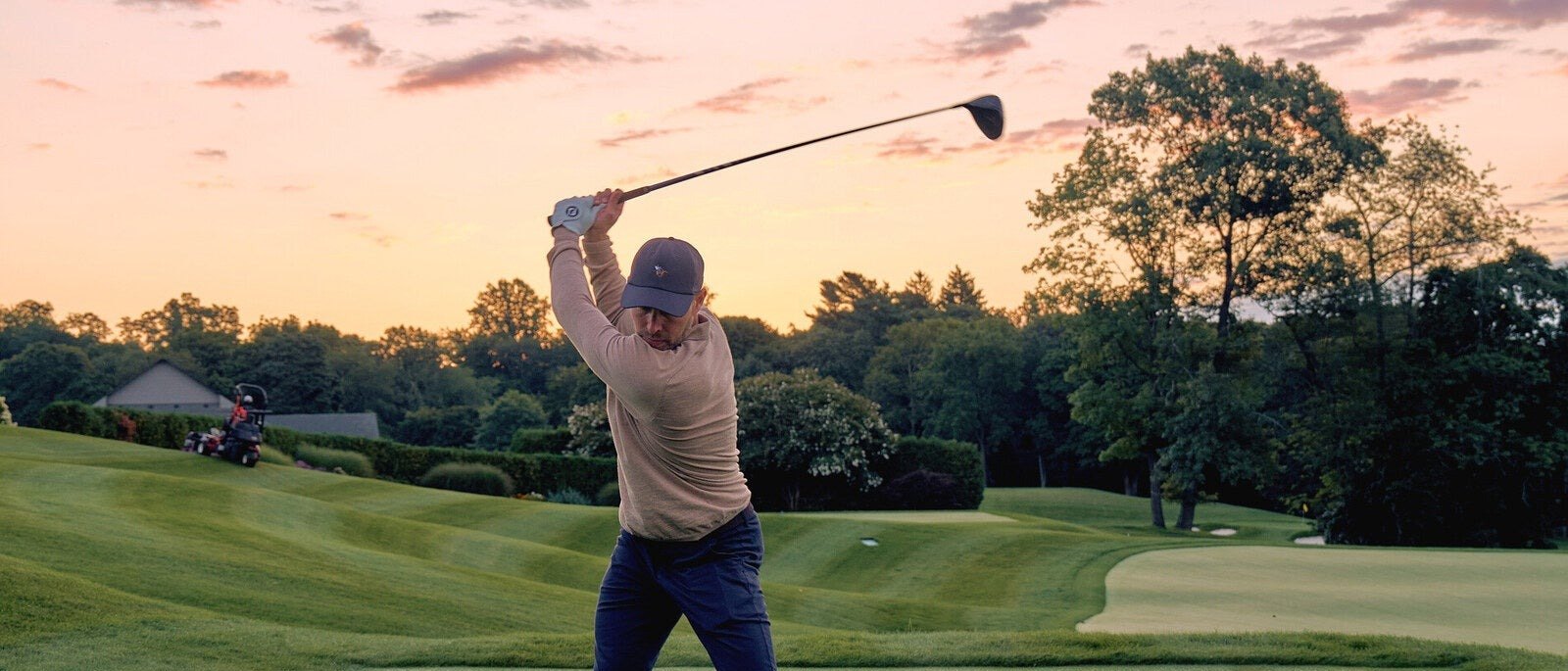 A golfer in an oatmeal sweater mid-swing on a tee at sunset