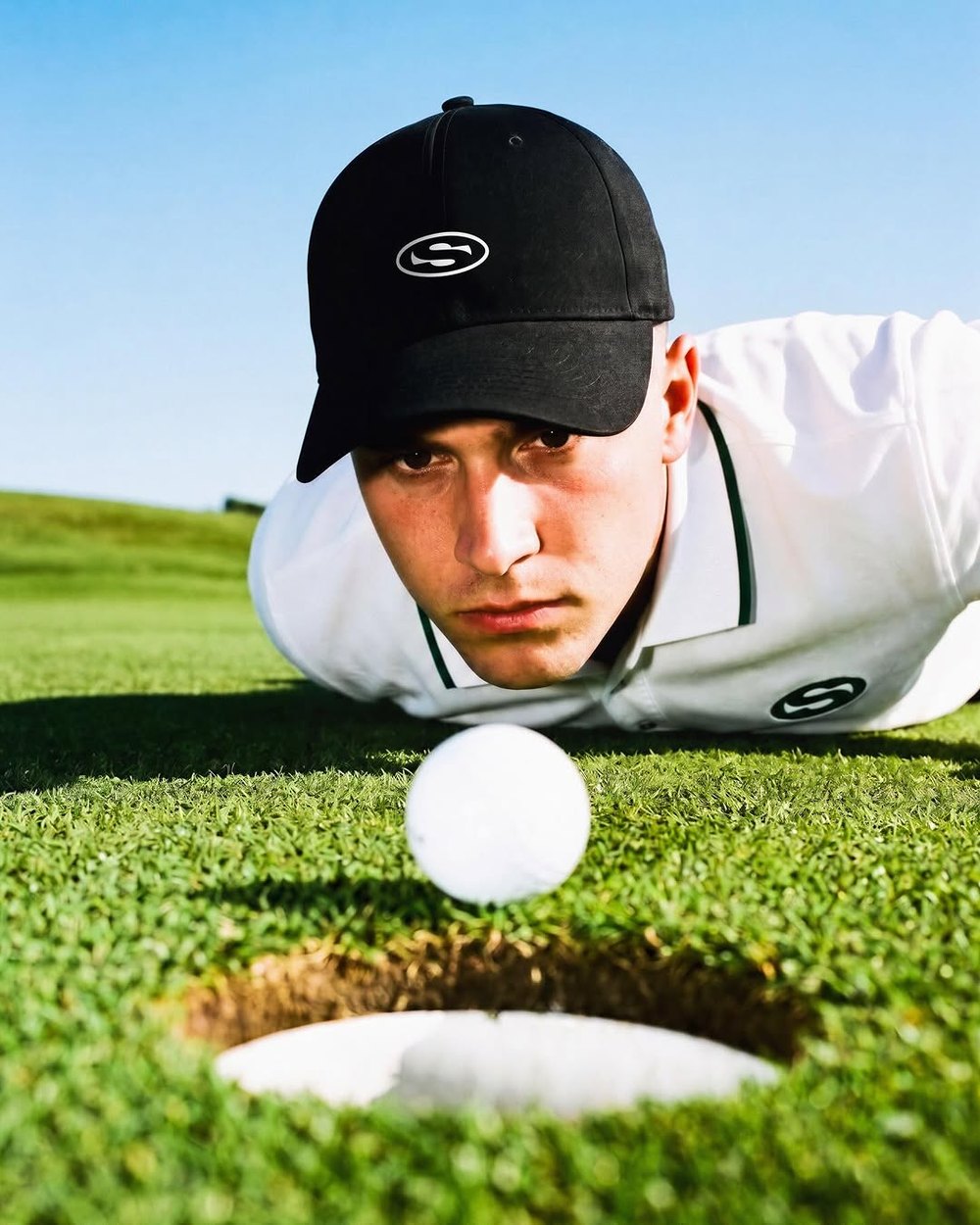 A golfer in the black Saha Logo Cap lying on the green beside the hole and ball