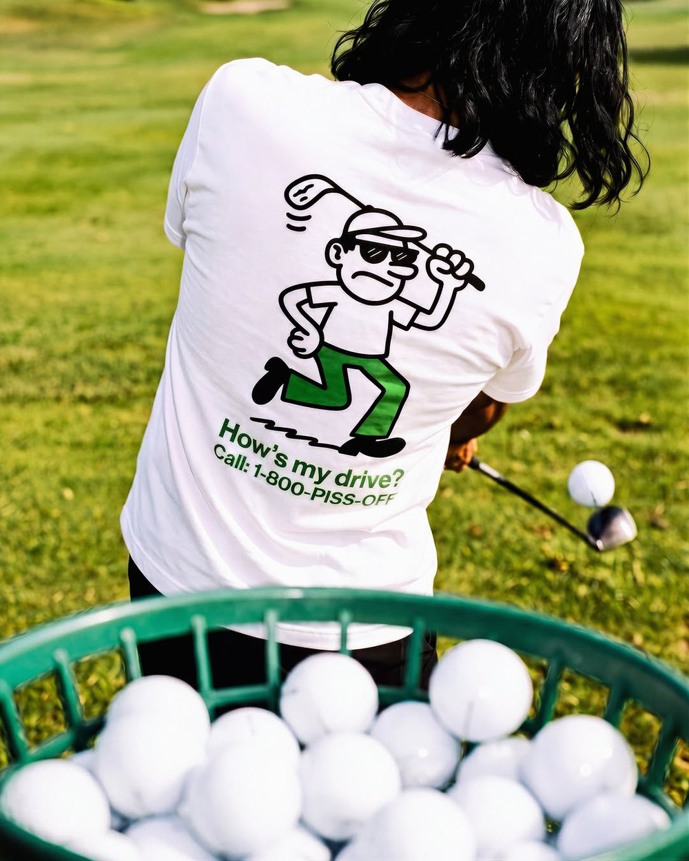 A golfer in the How’s My Drive Tee seen from behind at a range ball basket
