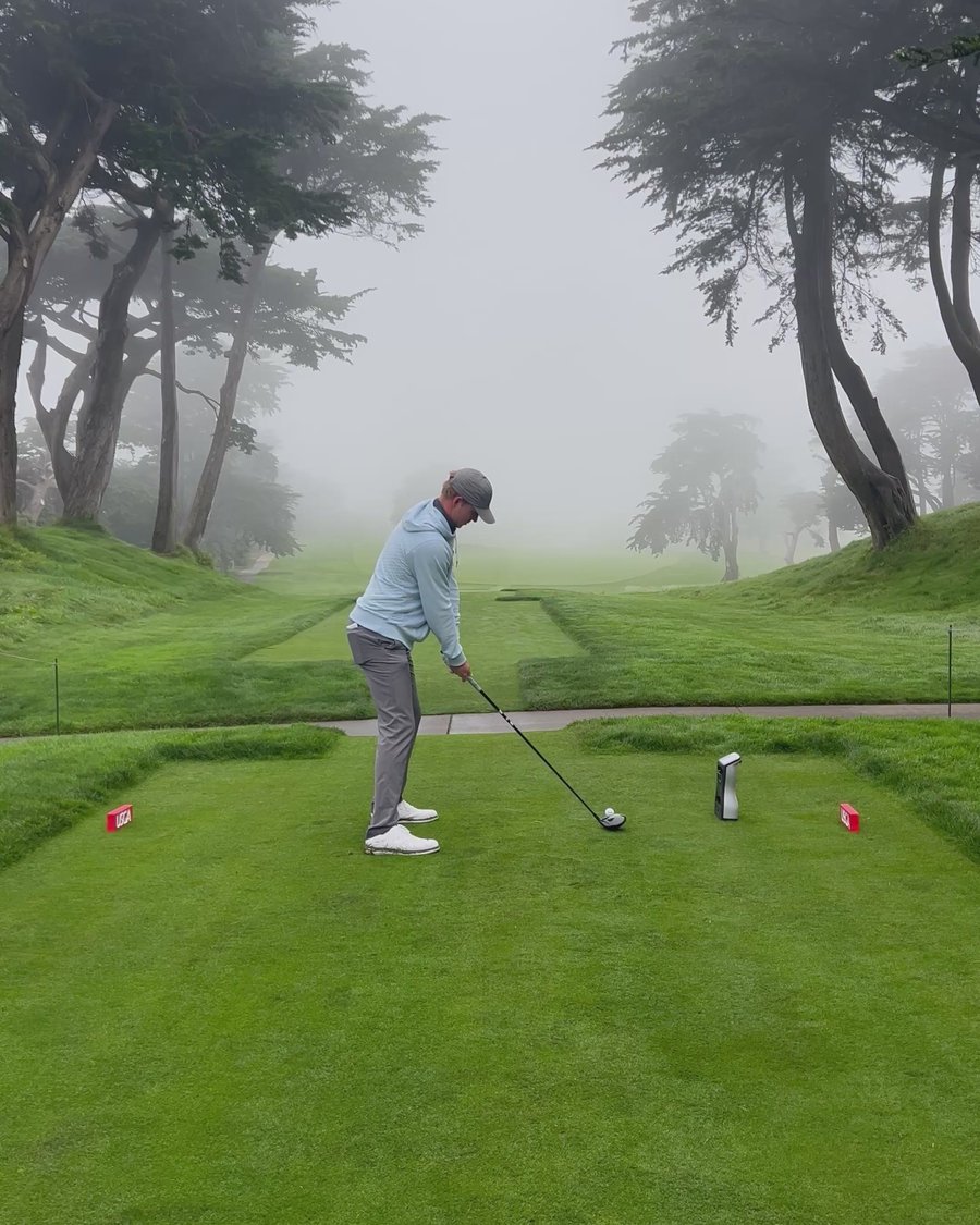 A golfer on a foggy tee box between cypress trees