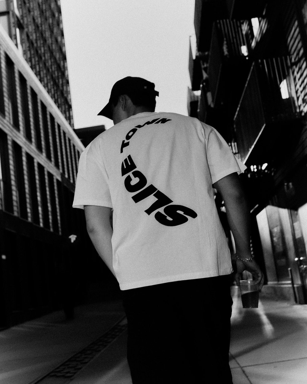 A man from behind in a white SLICE TOWN tee with the sliced logo on the back, between tall buildings