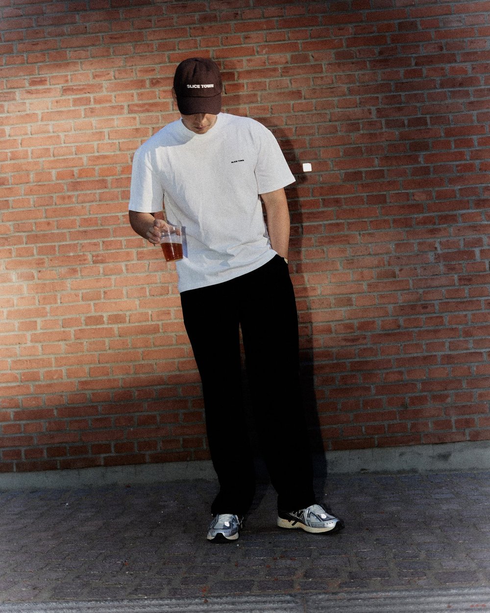 A man in a white tee and black trousers leaning on a brick wall with a pint