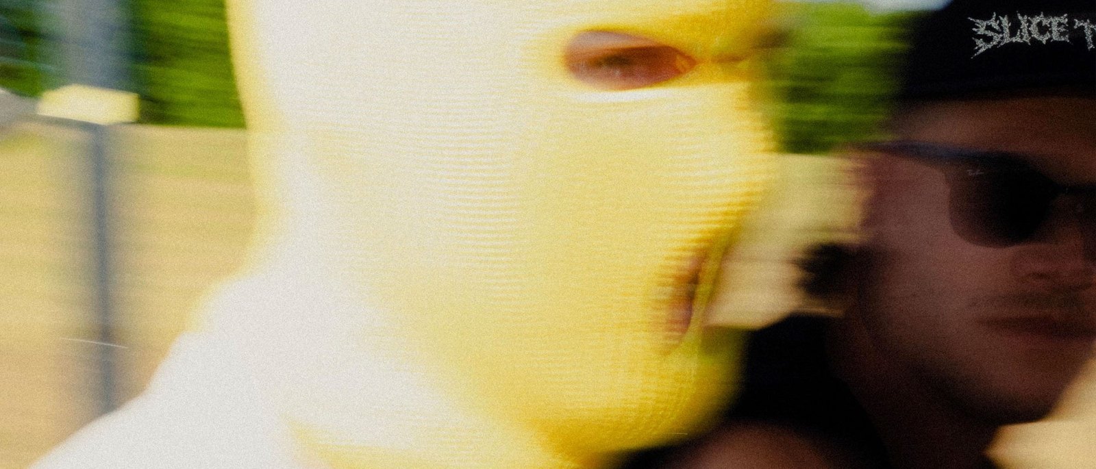 A golfer in a yellow knitted SLICE TOWN balaclava, blurred in motion, beside a friend in sunglasses and a SLICE TOWN cap