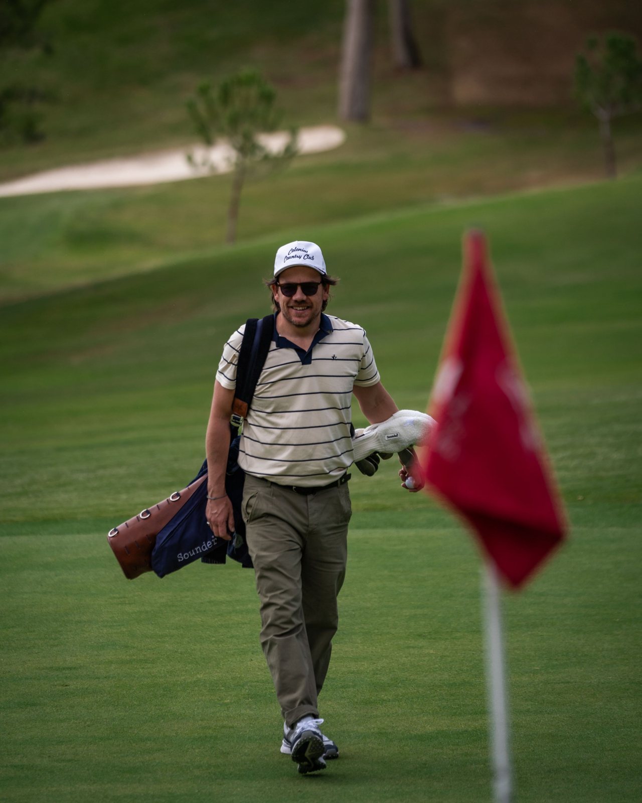 A golfer in a striped polo carrying a bag towards a flag on a course in Spain