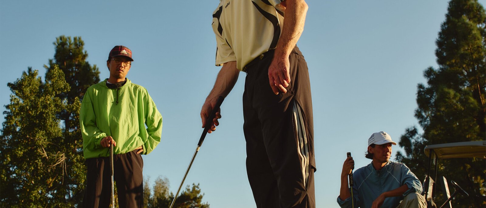 A golfer mid-swing in pleated trousers and an open camp shirt, a second figure in neon behind &mdash; Metalwood Studio campaign